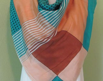 May include: A geometric patterned scarf in shades of peach, teal, brown, and white. The scarf is folded and draped over a white surface.