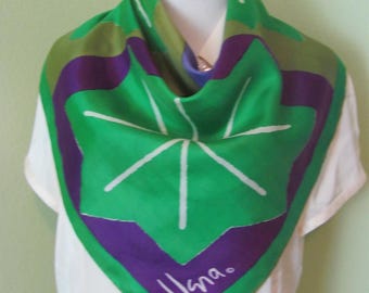 May include: A square silk scarf featuring a vibrant green base with bold purple geometric shapes and white line accents. The scarf displays a central star-like pattern and the white text "Vana." printed in a script font near the bottom corner.