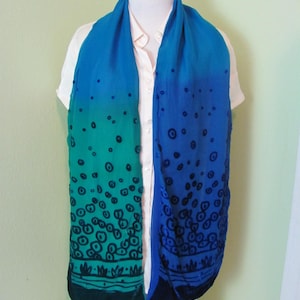 May include: A blue and green scarf with a unique design. The scarf features a gradient of colors, transitioning from blue to green. The design includes black circles and a dark green border at the bottom. The scarf is draped over a white shirt.