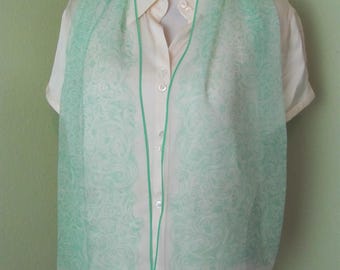 May include: A sheer, mint green scarf with a delicate, swirling pattern and a matching green border. The scarf is draped over a cream-colored button-down shirt, creating a layered effect. The scarf's design adds a touch of elegance to the outfit.