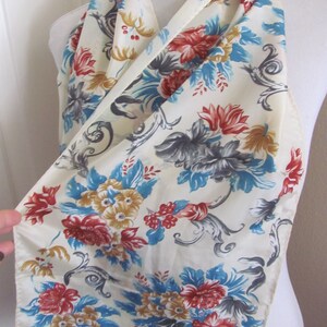 Pode incluir: Um lenço de seda branco com um padrão floral em tons de vermelho, azul e dourado. O padrão apresenta flores e folhas estilizadas sobre um fundo creme.