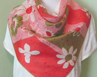 May include: A square silk scarf featuring a vibrant floral pattern in shades of coral, pink, and olive green. The design includes stylized white flowers and scattered red dots on a multi-colored background. The signature Ugra is printed in white.