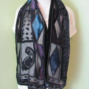 May include: A black sheer scarf featuring a patchwork design of velvet diamonds and paisley motifs in shades of blue and purple. The scarf is accented with thin white dotted lines and black velvet borders, creating a structured geometric pattern.