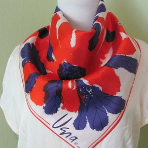 Peut inclure: Un foulard carré en soie avec un motif floral rouge, bleu et noir sur fond blanc. Le foulard est présenté sur un mannequin. La marque "Vana" est imprimée en cursive dans le coin.