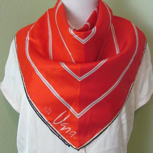 Peut inclure: Un foulard en soie orange vif avec un motif géométrique. Le foulard présente des accents blancs et noirs, avec le mot "Una" en écriture cursive. Le foulard est porté sur une chemise blanche.