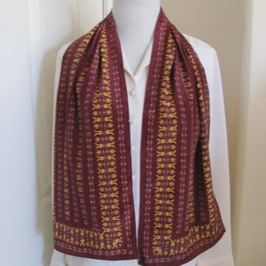 May include: A maroon silk scarf with a gold and white geometric pattern. The scarf is draped around a white shirt on a mannequin.