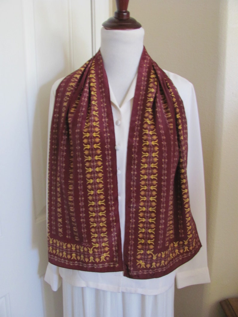 May include: A maroon silk scarf with a gold and white geometric pattern. The scarf is draped around a white shirt on a mannequin.