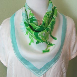 Peut inclure: Un foulard carré en soie avec un fond blanc et un motif floral vert et bleu. Le foulard a une bordure bleu clair et la signature "Una-is" dans le coin. Le foulard est drapé autour du cou d'un mannequin.