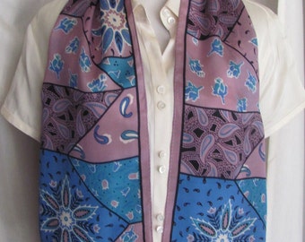May include: A purple, blue, and white patterned silk scarf with a paisley design. The scarf is folded in half and draped over a white shirt.