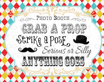 May include: A white sign with black text that says "Photo Booth. Grab a Prop. Strike a Pose. Serious or Silly. Anything Goes." The sign has a decorative border and is set against a background of red, yellow, and blue diamond shapes.