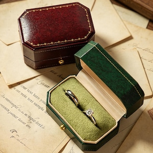 Puede incluir: Dos joyeros octogonales, uno burdeos y otro verde, se encuentran sobre un fondo de papel vintage. El joyero burdeos está cerrado, mientras que el joyero verde está abierto, mostrando dos anillos en un interior de terciopelo verde. Acentos dorados adornan los joyeros.
