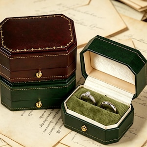 Puede incluir: Tres joyeros octogonales apilados. La caja superior es de color burdeos con ribete dorado, las cajas del medio e inferior son verdes. La caja verde abierta muestra dos anillos sobre un interior de terciopelo verde.