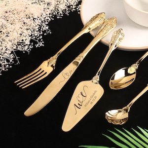 Könnte beinhalten: Eine Sammlung goldfarbener Servierutensilien, darunter eine Gabel, ein Messer, ein Tortenheber, Löffel und eine Kelle. Die Utensilien haben verzierte Griffe und sind auf einer schwarzen Oberfläche angeordnet. Der Tortenheber ist mit Initialen und einem Datum graviert.