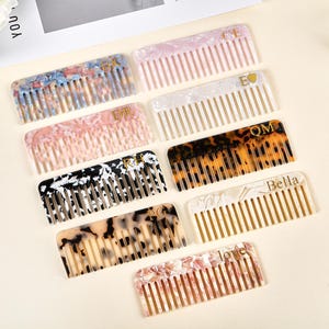 Könnte beinhalten: Eine Sammlung dekorativer Haarkämme in verschiedenen Farben und Mustern. Die Kämme weisen Marmor- und Schildpatt-Designs auf, einige sind mit Initialen oder Namen wie "Bella" und "love" personalisiert.