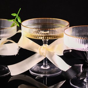 May include: Three clear champagne coupe glasses with gold rims and vertical ribbed detailing. Each glass is tied with a ribbon. One glass has a green stem. The glasses are on a black surface.