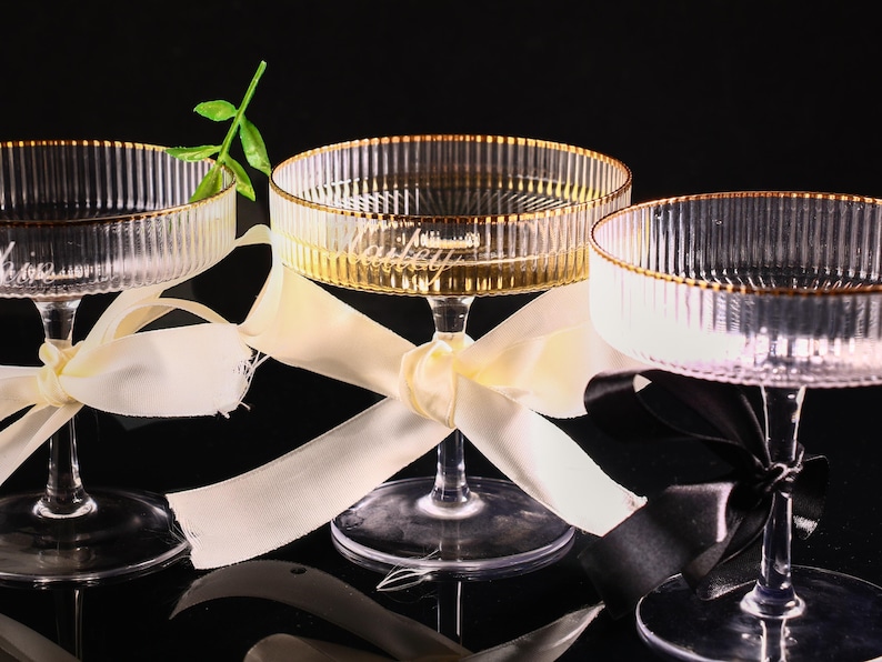 May include: Three clear champagne coupe glasses with gold rims and vertical ribbed detailing. Each glass is tied with a ribbon. One glass has a green stem. The glasses are on a black surface.