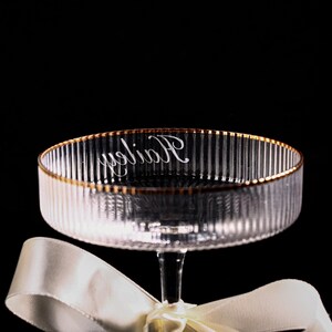 May include: Clear glass champagne coupe with vertical ribbed detailing and a gold rim. The word "Holiday" is written in cursive on the inside of the bowl. A white ribbon is tied around the stem.