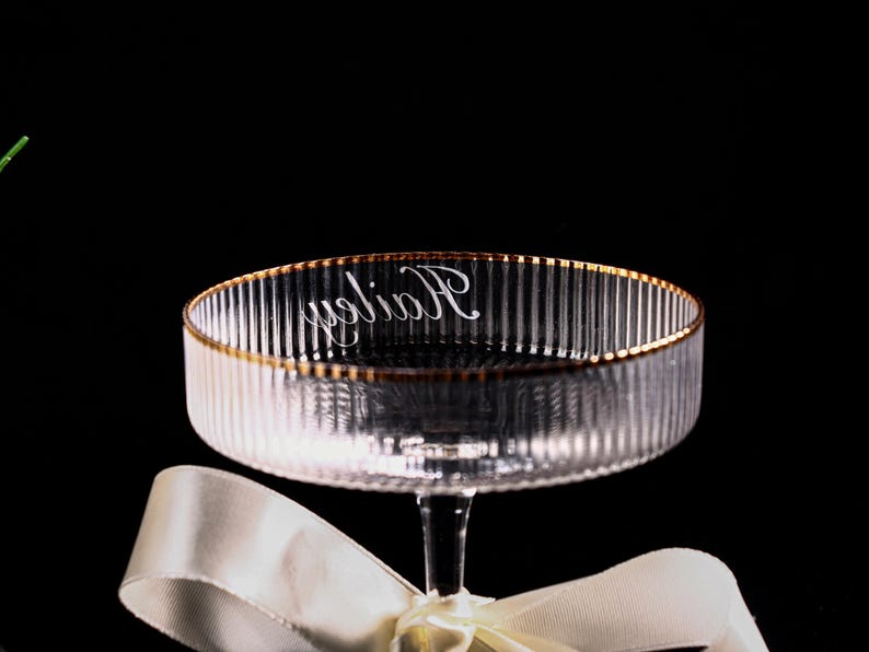 May include: Clear glass champagne coupe with vertical ribbed detailing and a gold rim. The word "Holiday" is written in cursive on the inside of the bowl. A white ribbon is tied around the stem.