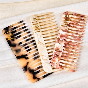 Könnte beinhalten: Drei dekorative Haarkämme. Einer ist schildpattfarben, einer cremefarben mit dem Aufdruck "Bella" und der dritte hat ein rosa-weißes Blumenmuster. Die Kämme liegen auf einer weißen Oberfläche.