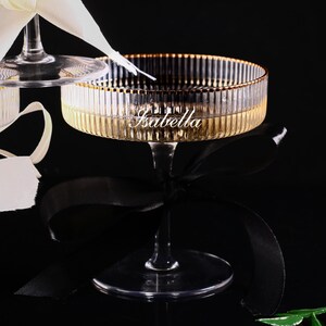 May include: Elegant champagne glasses with a gold rim and a ribbed design. The word "Isabella" is inscribed on the glass. The glasses are adorned with white and black ribbons, set against a dark background.
