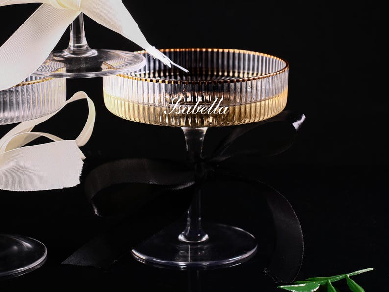 May include: Elegant champagne glasses with a gold rim and a ribbed design. The word "Isabella" is inscribed on the glass. The glasses are adorned with white and black ribbons, set against a dark background.