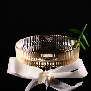 May include: A clear glass bowl with a ribbed design and gold trim, resting on a white ribbon. The bowl has the word "Radion" etched on the side. A sprig of green leaves is placed on the side of the bowl.