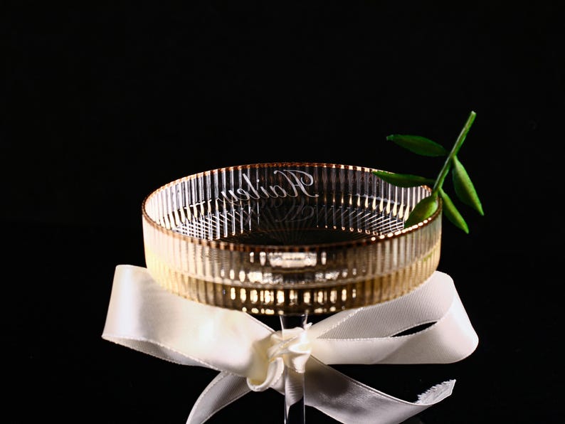 May include: A clear glass bowl with a ribbed design and gold trim, resting on a white ribbon. The bowl has the word "Radion" etched on the side. A sprig of green leaves is placed on the side of the bowl.