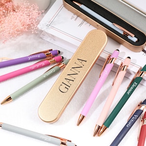 Puede incluir: Un estuche de metal dorado para bolígrafos con el nombre "Gianna" grabado en él. El estuche está abierto y se ven varios bolígrafos con cuerpos de diferentes colores y detalles en oro rosa. Los bolígrafos están personalizados con nombres como "Victoria", "Eleanor", "Camilla", "Gabrielle" y "Gianna".