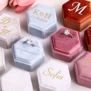 Op de afbeelding: Een verzameling gepersonaliseerde zeshoekige fluwelen ringendoosjes in verschillende kleuren, waaronder lichtblauw, roze en oranje. Sommige doosjes hebben initialen, data of namen in gouden letters. Verschillende doosjes bevatten diamanten verlovingsringen.