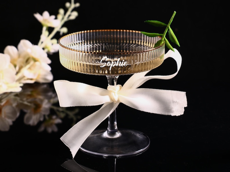 May include: A champagne coupe with a ribbed bowl and gold rim, tied with a white ribbon. The glass is engraved with the name "Sophie". A sprig of greenery and white flowers are in the background.