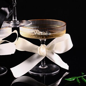 May include: Elegant champagne glasses with gold rims and the name "Victoria" etched on the side. Each glass is adorned with a satin ribbon bow. The glasses are set against a dark background, creating a sophisticated aesthetic.