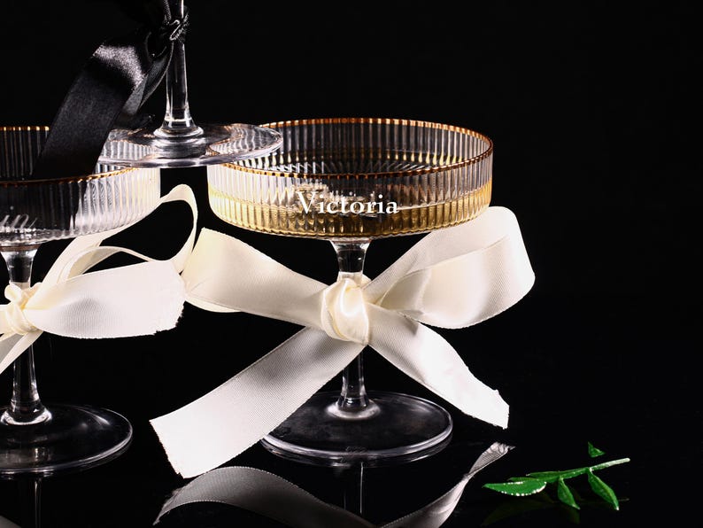 May include: Elegant champagne glasses with gold rims and the name "Victoria" etched on the side. Each glass is adorned with a satin ribbon bow. The glasses are set against a dark background, creating a sophisticated aesthetic.