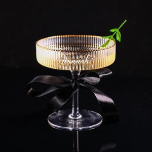 May include: A clear glass cocktail glass with a gold rim and the name "Hannah" etched on the side. A black ribbon is tied around the stem. A small green sprig sits on the rim. The glass is on a black surface.