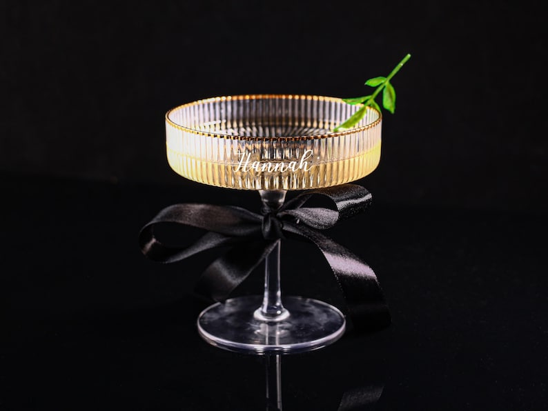May include: A clear glass cocktail glass with a gold rim and the name "Hannah" etched on the side. A black ribbon is tied around the stem. A small green sprig sits on the rim. The glass is on a black surface.