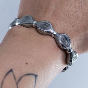 Op de afbeelding: Een zilveren armband met een reeks getextureerde, ovaalvormige schakels. De armband wordt om een pols gedragen, met een bloementattoo zichtbaar op de arm. De schakels van de armband hebben een geribbeld patroon.