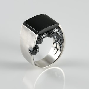 May include: A silver ring with a large, square black onyx stone. The ring features a detailed, textured design on the sides. The ring is displayed on a white surface, showcasing its reflective qualities. A modern piece of jewelry.