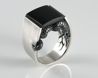Puede incluir: Un anillo de sello de plata con una piedra negra rectangular plana en la parte superior. La banda tiene un acabado de plata cepillada e incorpora un motivo de serpiente oscuro, texturizado y detallado que envuelve el lateral del anillo.
