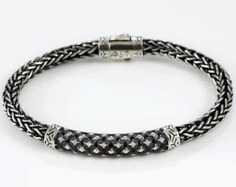 May include: A silver bracelet with a woven chain design. The bracelet features a central cylindrical section with a geometric pattern and decorative end caps. The clasp is also silver and has an intricate design.