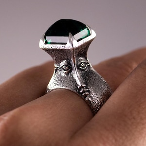 May include: A silver ring featuring a large, emerald-cut, dark green gemstone. The ring's band has a textured design with two stylized faces. The ring is shown on a hand, highlighting its unique design and colour.