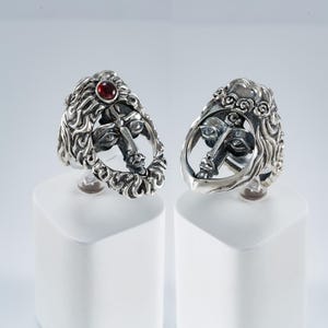 Könnte beinhalten: Zwei silberne Ringe mit Gesichtsdesign. Ein Ring hat einen roten Edelstein. Die Ringe werden auf weißen Sockeln vor hellem Hintergrund präsentiert. Das Gesichtsdesign umfasst detaillierte Haare und Gesichtszüge.