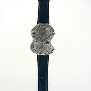 May include: A unique wristwatch with a silver-toned, figure-eight shaped face and a navy blue suede strap. The watch face features intricate detailing and two separate dials with blue hands. The buckle and face are adorned with decorative patterns.