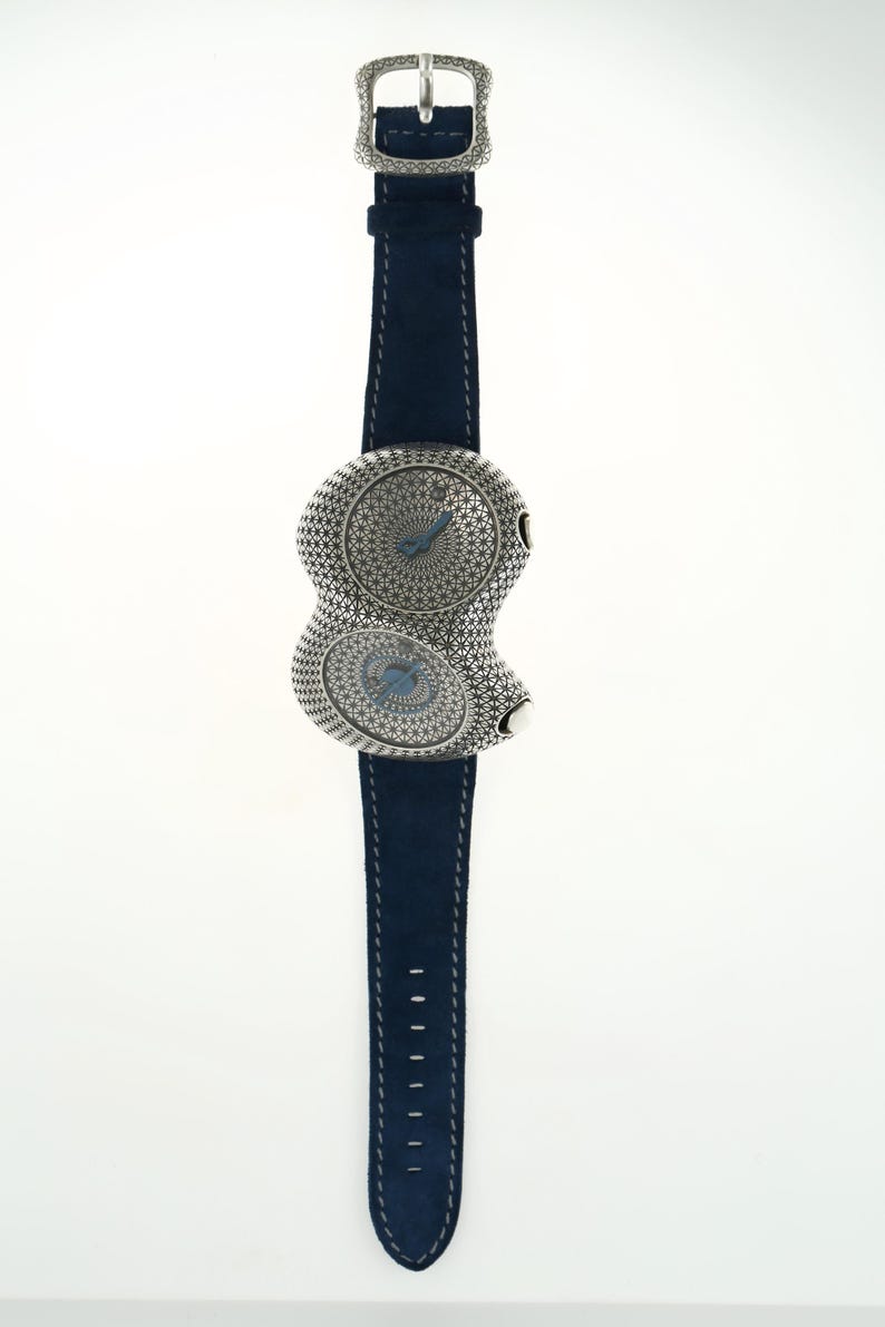 May include: A unique wristwatch with a silver-toned, figure-eight shaped face and a navy blue suede strap. The watch face features intricate detailing and two separate dials with blue hands. The buckle and face are adorned with decorative patterns.