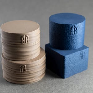 May include: Two beige cylindrical objects with horizontal ridges stacked vertically, alongside a blue cylindrical object resting on a blue square object. Each of the four items features a small, embossed logo consisting of two vertical lines.