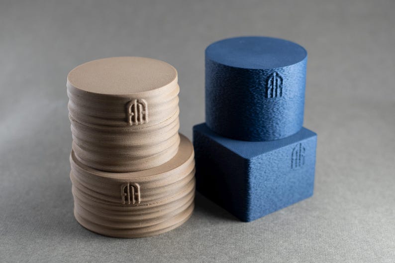 May include: Two beige cylindrical objects with horizontal ridges stacked vertically, alongside a blue cylindrical object resting on a blue square object. Each of the four items features a small, embossed logo consisting of two vertical lines.