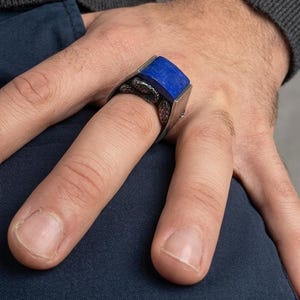 May include: A silver ring featuring a rectangular, deep blue lapis lazuli stone set horizontally. The band of the ring is designed with a textured, dark metallic snake motif that wraps around the finger, providing a detailed contrast to the smooth, vibrant blue stone.