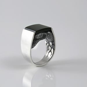 May include: A silver ring featuring a smooth, rectangular black stone set at the top. The band is crafted with a textured, scale-like pattern that wraps around the sides, transitioning into a polished, solid silver finish on the outer shank.