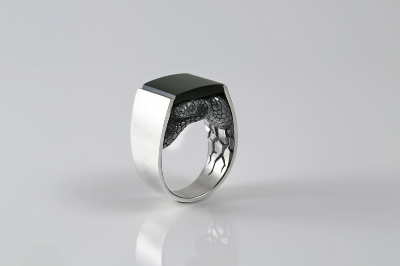 May include: A silver ring featuring a smooth, rectangular black stone set at the top. The band is crafted with a textured, scale-like pattern that wraps around the sides, transitioning into a polished, solid silver finish on the outer shank.