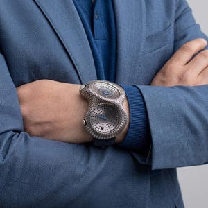 May include: A unique, silver-toned wristwatch with a distinctive perforated design. The watch features two circular faces and a dark blue leather band. The watch is worn on a wrist, with the person wearing a blue blazer and sweater.