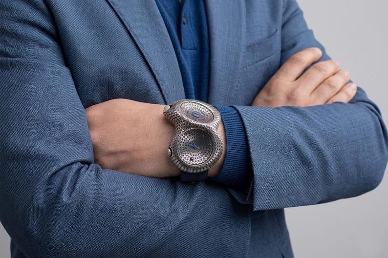 May include: A unique, silver-toned wristwatch with a distinctive perforated design. The watch features two circular faces and a dark blue leather band. The watch is worn on a wrist, with the person wearing a blue blazer and sweater.