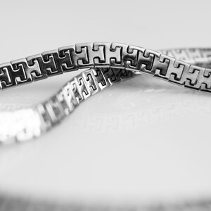 May include: A silver-toned metal chain bracelet featuring a repeating geometric H-shaped link pattern. The chain is flexible and displays a polished, reflective surface with a series of interconnected, square-edged links that form a continuous band.